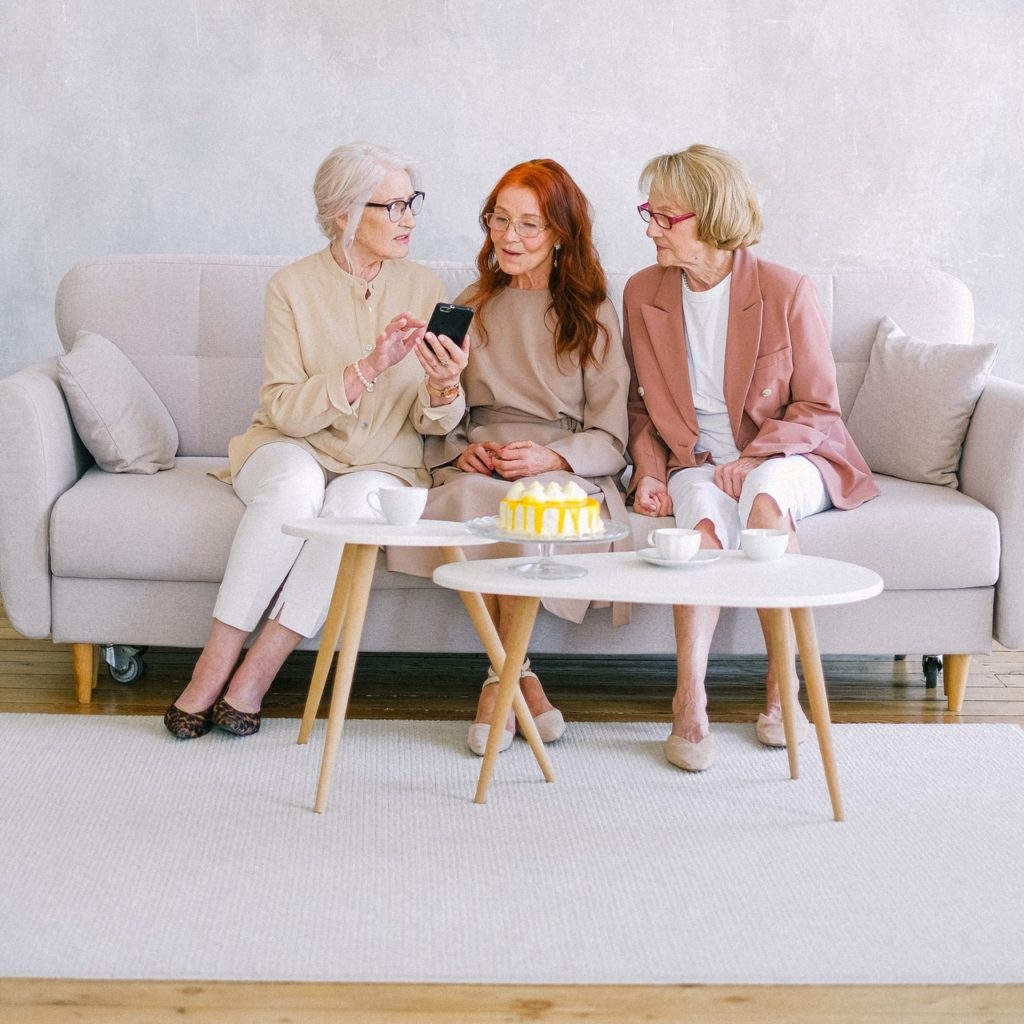 3 dames âgées assises sur un canapé gris discutant et l'une d'entre elles montre quelque chose sur son smartphone